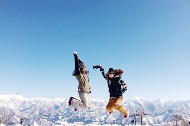 雪山の山頂で、スノーボードウェアを着た女性二人が、青空に向かって元気いっぱいにジャンプしている。最高のロケーションで、冬の思い出を作る一枚。