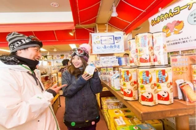 スキー場でのお土産物屋。スキーウェアを着たカップルが、お土産を選びながら笑顔で会話をしている。