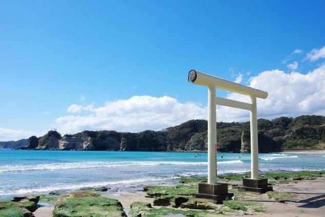 青い空とエメラルドグリーンの海、白い鳥居が立つビーチ。サーフィンを楽しむ人々の姿も見え、リゾートバイトで海辺での仕事やマリンスポーツに興味がある方に魅力的な画像。