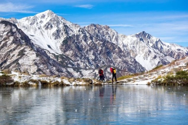 雪を抱く山々と氷を張った湖。登山を楽しむ2人の人物が映る壮大な絶景のイメージ画像。リゾートバイトの休日を満喫。