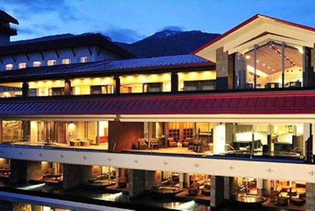 リゾートバイトの勤務先ホテルの外観の夜の様子。暖かくライトアップされた窓やバルコニー、その奥に見える山々が印象的。