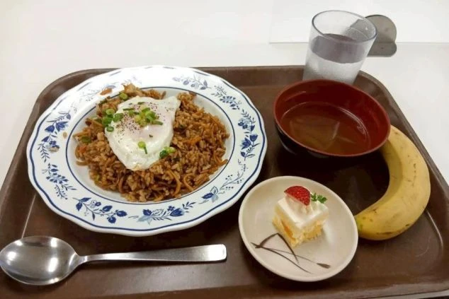 ボリューム満点のまかないの画像。目玉焼きがのったそばめし、スープ、バナナ、ミニケーキの豪華なセット。