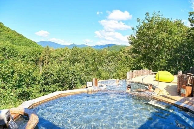 露天風呂でくつろぐ人々の画像。青い空と緑豊かな山々を背景に、広々とした温泉が広がる。リゾートバイトの勤務後にリラックスできる温泉施設。