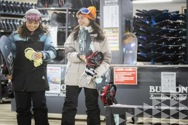 スキーウェアを着た女性たちが、スノーボードレンタル店でボードを選んでいる画像。リゾートバイトの休日にスノボを楽しむイメージ。
