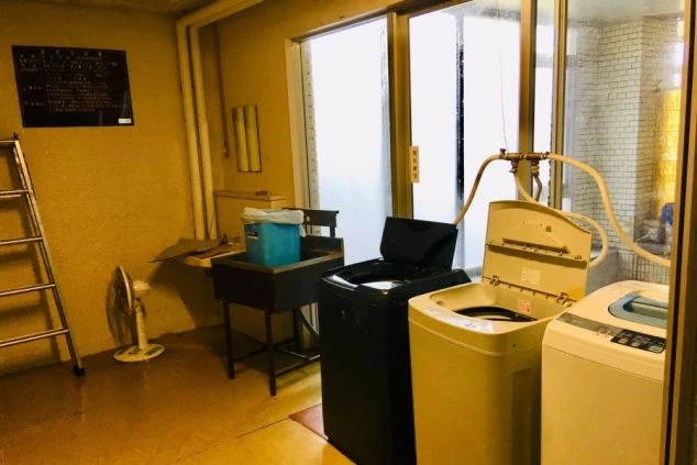 寮のランドリー。洗濯機とシンクが並び、扇風機や脚立が置かれた共用空間が映る。