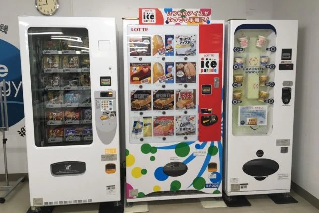 寮の自動販売機。アイス、お菓子、カップ麺が並び、手軽に買える設備が映る。