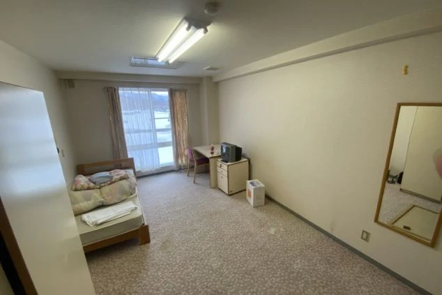 寮の個室。ベッド、テレビ、机、椅子、クローゼット、鏡が映る部屋の様子。