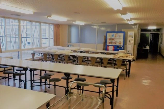 リゾートバイト寮の食堂の画像。テーブルと椅子、布団が置かれ、自動販売機がある共用スペースが映る。