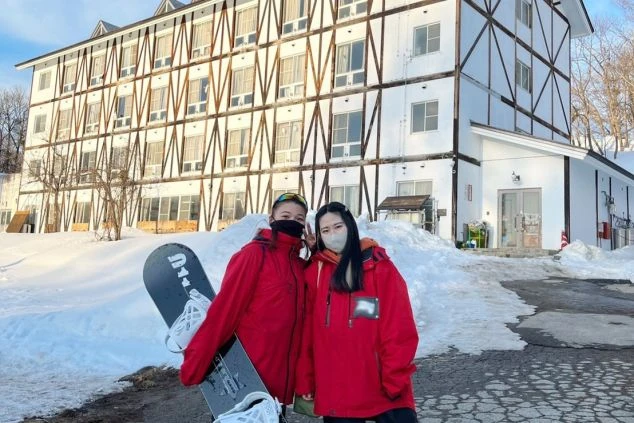 ワクトリでリゾートバイトをしたスタッフが撮影した、雪が積もるロッジの前で、スノーボードを持つ女性2人が笑顔でポーズをとるイメージ画像。リゾートバイトの仲間との休日を満喫。
