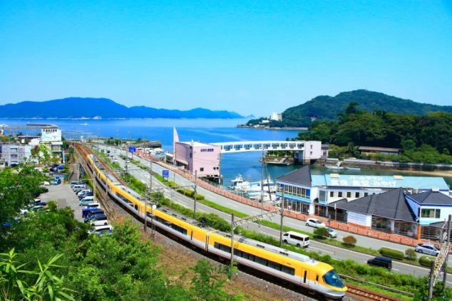 電車が走る線路と、その奥に広がる港と海。青い空と海の間に山々が連なる、開放的な景色。