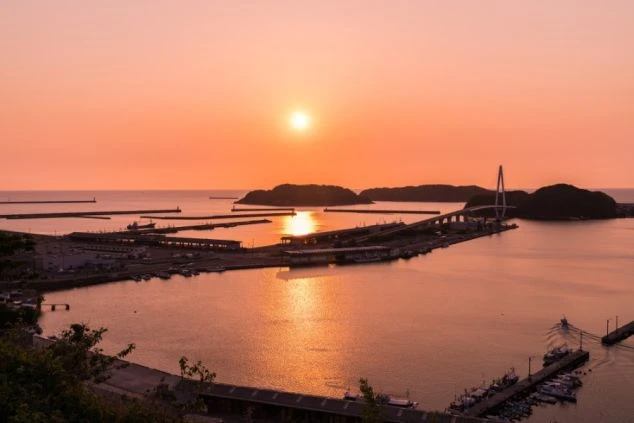 日の出か日の入りの時刻、オレンジ色に染まる港。海に架かる橋がライトアップされ、幻想的な雰囲気を醸し出す。