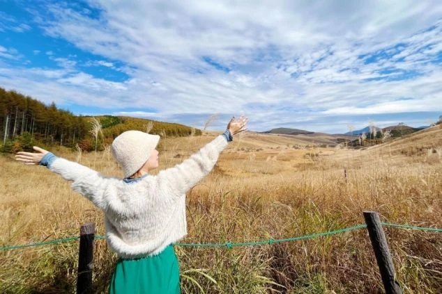 白樺湖周辺の草原で、ススキが揺れる中で両手を広げてリフレッシュする女性の後ろ姿。リゾートバイトの仕事の合間に、自然の中で癒やしの時間を過ごしたい方に魅力的なワクトリスタッフが撮影したオリジナル画像。
