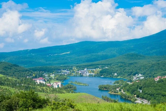 長野県白樺湖の夏の風景。緑の山々に囲まれた湖畔にホテルや建物が並び、リゾートバイトで湖畔のリゾート施設やレジャー関連の仕事を探している方に訴求する画像。