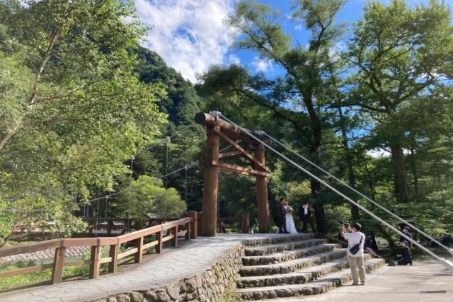 緑の山と川に架かるつり橋。橋の上ではウェディングドレスとタキシードを着たカップルが記念撮影をしており、リゾートバイトで結婚式場やホテルでの仕事に興味がある方に訴求する画像。