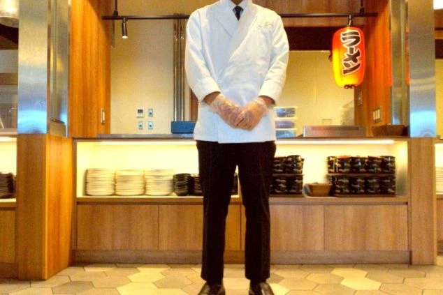 白いコックコートを着用し、調理場のカウンターの前に立つ男性。リゾートバイトで調理スタッフやキッチン業務に興味がある方に、仕事内容をイメージさせる画像。