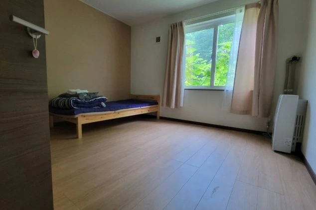 寮の個室。木目調の床にベッド、窓、暖房器具が映るシンプルな部屋の様子。