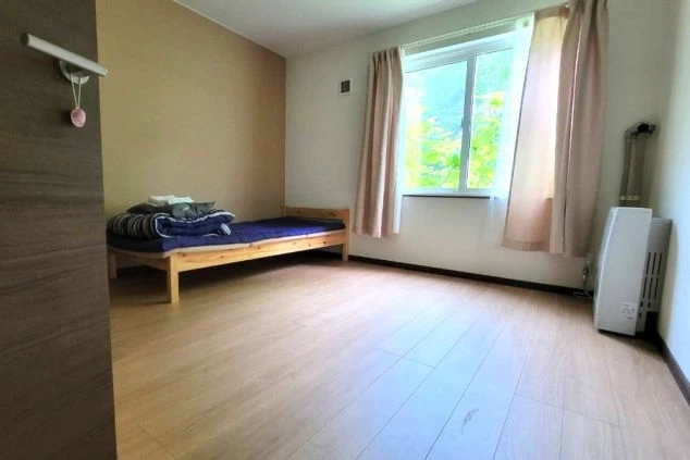 寮の個室。木目調の床にベッド、窓、暖房器具が映るシンプルな部屋の様子。