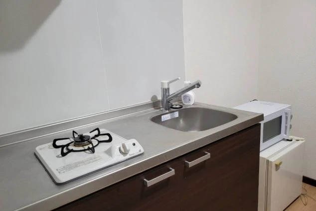 寮のミニキッチン。シンク、コンロ、レンジ、冷蔵庫が備え付けられたコンパクトな空間が映る。