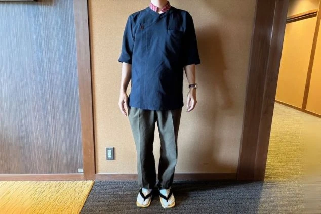 濃紺の半袖シャツにカーキ色のズボンを着用し、足元には草履を履いた男性スタッフの画像。和風の落ち着いたデザインで、旅館や和食レストランでの仕事内容を表す、サービススタッフの制服画像。