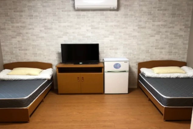 寮の部屋。ベッド2台とテレビ、冷蔵庫、エアコンが映る部屋の様子。