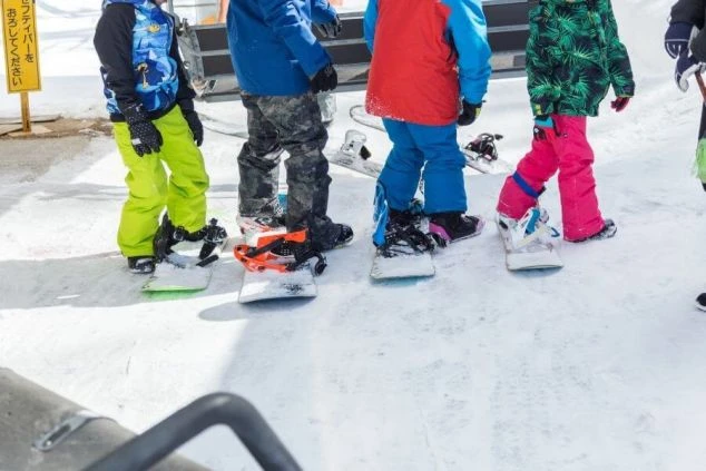 カラフルなスノーボードウェアを着た子供たちが、スノーボードを装着して雪の上に並んでいる様子。スキー場のキッズスクールやレンタルスタッフなど、子供と関わるリゾートバイトに興味がある方に訴求する画像。