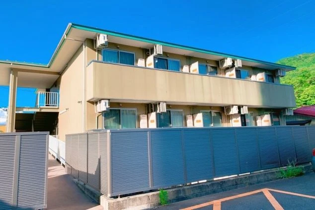 寮の外観。薄いベージュ色の2階建てアパート風の建物で、各部屋の窓の外にエアコンの室外機が多数並んでいる。手前には目隠しのフェンスがある。