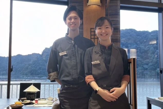 窓から海が見えるレストランで、和風モダンの制服を着用した男女のスタッフが笑顔で立つ写真。リゾートバイトでレストランサービスやフロント業務に興味がある方に訴求する画像。