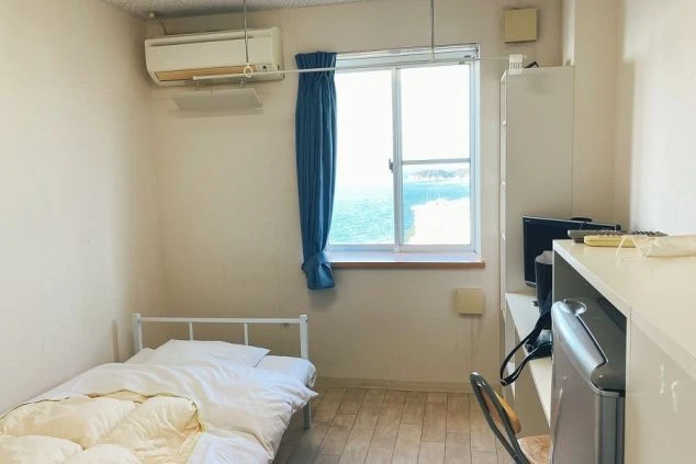 リゾートバイトの寮の部屋の画像。窓から海が見え、ベッド、テレビ、冷蔵庫、エアコンが完備されている。海が見える寮は、リゾートバイトの魅力を高める。