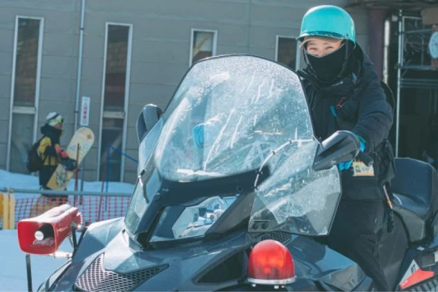 ヘルメットをかぶり、スノーモービルに乗ったパトロール隊員らしき人物。背景には建物とスノーボードを持った別の人物が見える。