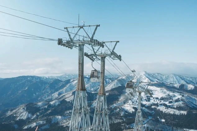 快晴の空を背景に、雪山の上をゴンドラリフトが通過している。遠くには雪をかぶった山々が連なっている。