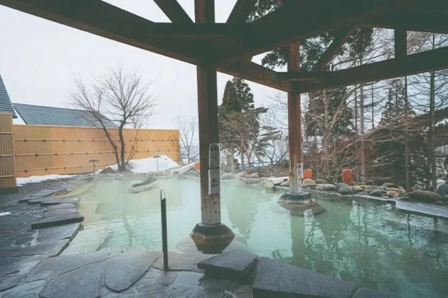 雪に囲まれた露天風呂。湯船からは湯気が立ち上り、周りには岩や木々、竹垣が見える。屋根付きの休憩スペースが写真の手前にある。