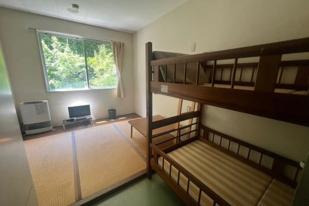 畳敷きのスペースと緑の床からなる、古い寮の部屋。窓の外には木々が見え、部屋にはテレビと二段ベッドがある。