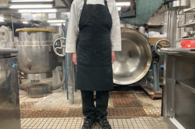 白いコックコートと黒いエプロン、黒いズボンを着用した男性。背景には大きな調理器具が見える。