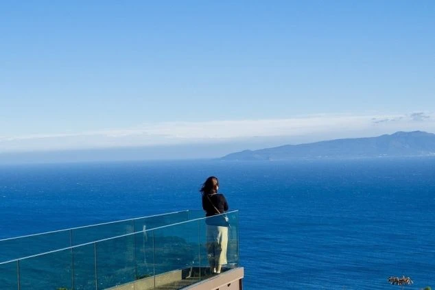 青い空と広大な海を、ガラス張りの展望台から眺める女性の後ろ姿。リゾートバイトで絶景の中で働き、仕事の合間にリフレッシュしたい方に魅力的な画像。
