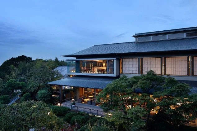 夕暮れ時、庭園の緑の中に佇む和風モダンの旅館。窓から温かい光が漏れ、リゾートバイトの勤務地をイメージさせる、落ち着いた雰囲気の外観の画像。
