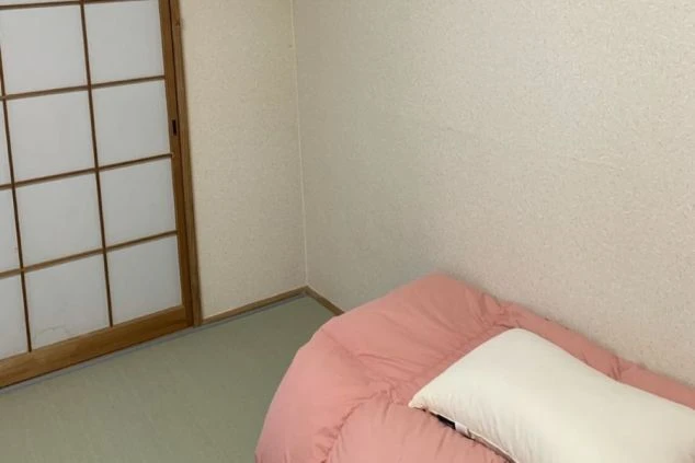 明るい部屋の一角を写した画像。左側には伝統的な障子または和紙のスクリーンが見え、右側には白い枕が置かれた薄いピンク色のベッドまたは布団が写っています。部屋の壁は薄い色のようです。