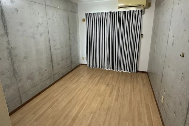 コンクリート打ちっぱなしの壁が特徴的な部屋。フローリングの床で、窓にはストライプ柄のカーテンがかかっています。