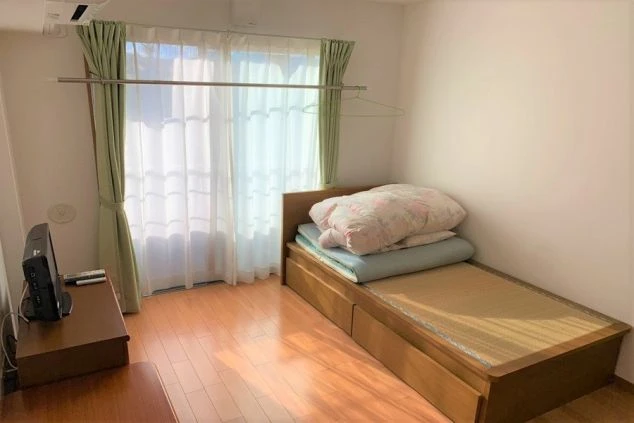 フローリングの床で、窓際に布団が積まれ、窓の横にはテレビが置いてあるリゾートバイトの個室寮の画像。