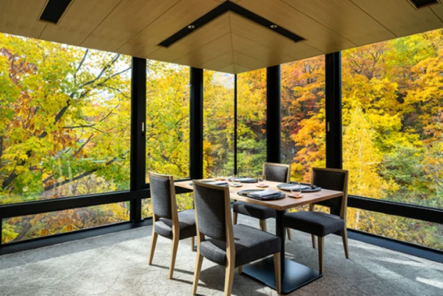 窓の外に紅葉した木々が広がるダイニングルーム。木製のテーブルと4脚の椅子が置かれている。