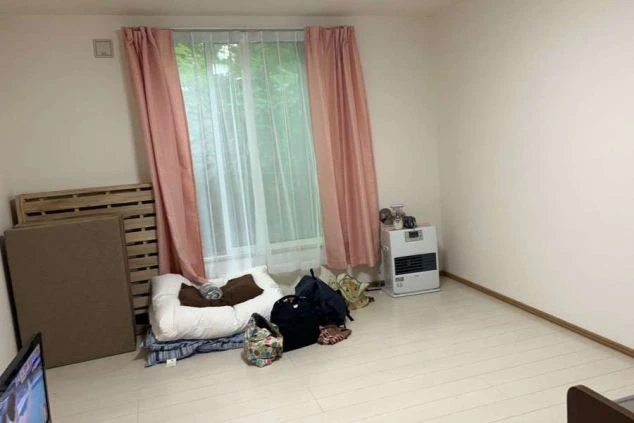 寮の個室。木製床に窓、ピンクのカーテン、暖房器具、扇風機が映る部屋の様子。