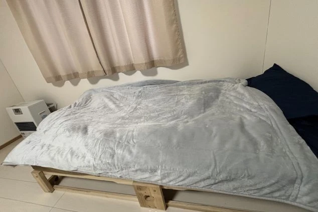 フローリングの床に布団が敷かれ、窓にはピンクのカーテンがかかっている。部屋の隅にはヒーターと荷物がある。