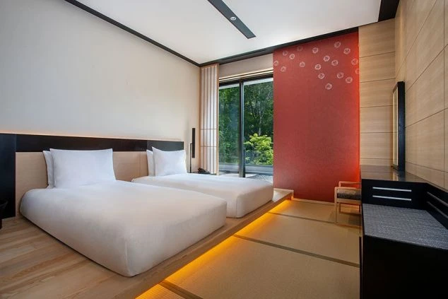 ツインベッドが置かれたモダンな和室。壁の一部は赤く、窓の外には緑の木々が見える。