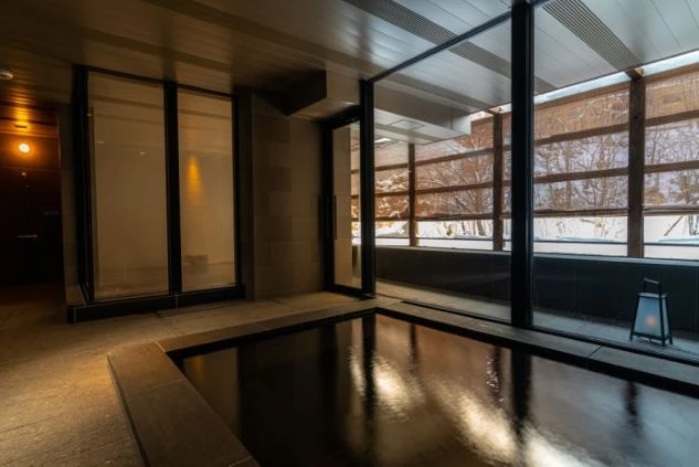 大きな窓から雪景色が見えるモダンな内風呂。浴槽には温泉が満たされており、湯気が立っている。