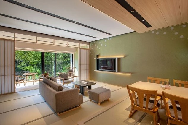 モダンな内装の和室のリビングルーム。畳敷きの床にソファとテーブルが置かれ、壁にはテレビがかけられている。窓からは緑豊かな景色が見える。