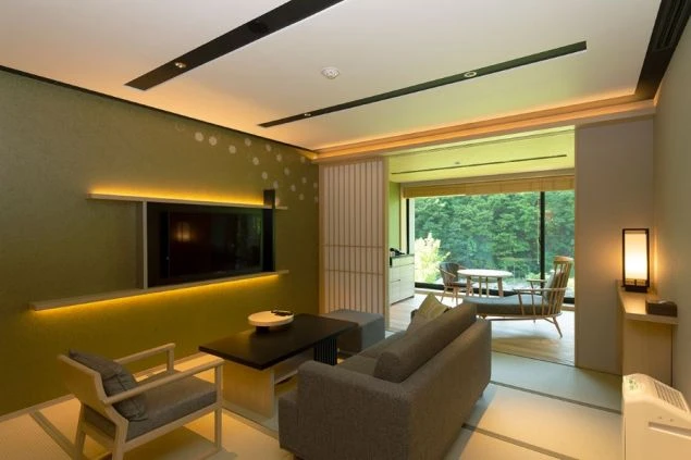 モダンな和室のダイニングとリビングルーム。畳敷きの床にはダイニングテーブルセットとソファが置かれ、壁にはテレビがかかっている。窓からは緑豊かな景色が見える。