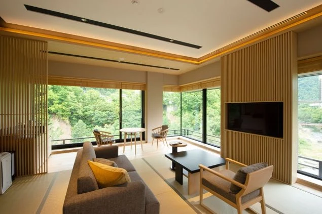 窓から緑の木々が見える、モダンな和室のリビング。畳敷きの床にソファや椅子、テーブル、テレビが置かれている。
