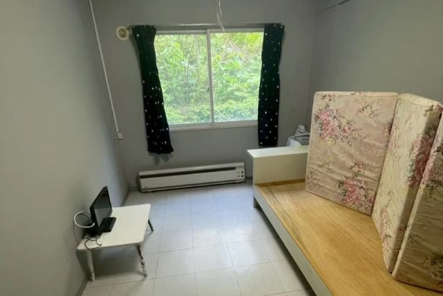 寮の居室。フローリングの床で、窓際に布団が敷かれている。部屋にはエアコンが設置され、テレビ台と小型テレビが見える。