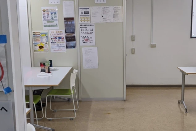 寮の食堂の掲示板。週替わりのメニュー表、営業時間、そしてステーキやセットメニューの告知ポスターなどが貼られ、下の台にはサンプル料理名(アジフライなど)の札が置かれている。