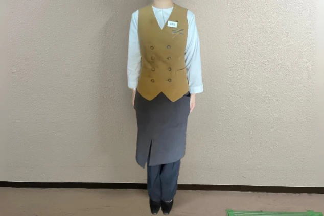 白い長袖シャツに茶色のダブルボタンベスト、黒いスカートとパンツを着用し、ハーフエプロンを重ねた女性料飲部スタッフの全身画像。
