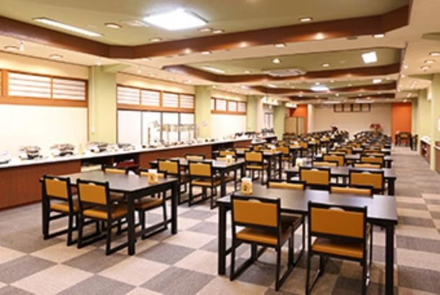 広々としたホテルのダイニングルーム。複数のテーブルと椅子が規則正しく並び、左側にはビュッフェ形式の料理が並んでいる。